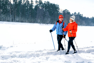 Smiling senior couple walking with nordic walking poles in snowy winter park. Elderly wife and husband doing healthy exercise outdoors. Active lifestyle concept.