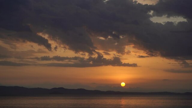 4k stock video footage of morning seascape of Greece. Cloudy dramatic sunrise sky with magic sun over dark silhouettes of mountains seen on horizon line, sunlight on calm surface of blue sea