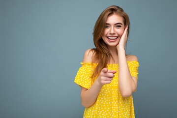 Attractive sexy smiling good looking young blonde woman wearing stylish yellow summer dress standing isolated over blue background communicating on mobile phone looking at camera and showing phone