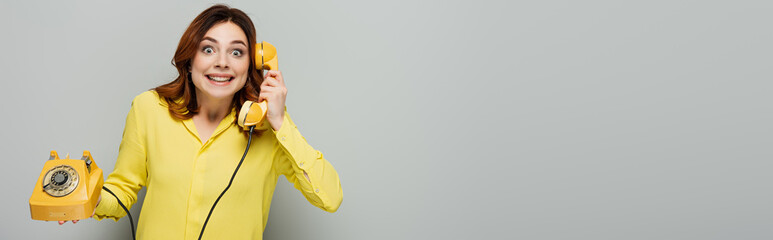 cheerful woman talking on vintage telephone while looking at camera on grey, banner