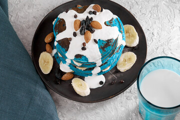 Vegan blue pancakes with sour cream and banana slices on a plate sprinkled with almonds, raisins, next to a glass of milk on a gray textured background