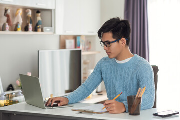 Side view of an Asian man working at a home desk using a tablet and taking notes.