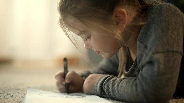 Little, happy preschool girl drawing with a marker in her notebook, smiling and laughing laying on the carpet. Artistic children and apartment concept.
