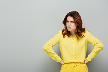 irritated woman looking away while standing with hands on hips on grey