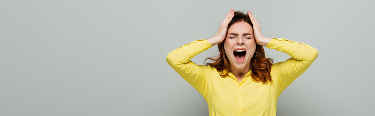 angry woman screaming while standing with closed eyes and touching head on grey, banner