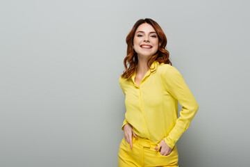 young curly woman in yellow shirt standing with hands in pockets on grey