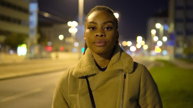 A young black woman looks seriously at the camera in a street in an urban area at night