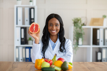Positive black nutrition expert holding grapefruit at desk in medical office