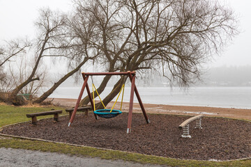 Children's playground with a swing on the bank of the dam at the frozen water level. There is fog in the background.