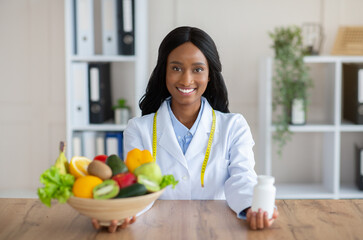 Black female dietitian holding bowl of fresh products and jar of tablets at table in clinic....