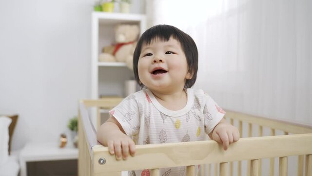 tilt up adorable asian baby is smiling at the distance and babbling while leaning on the wooden crib playpen in the bedroom at home.