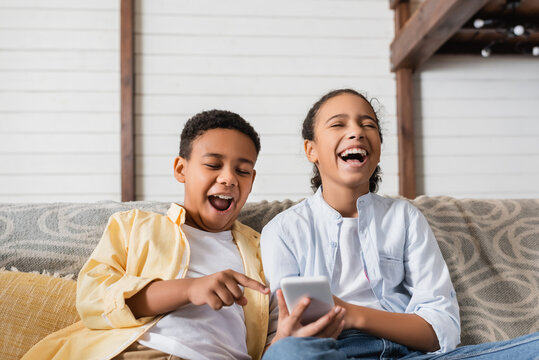 excited african american kids using smartphone on sofa
