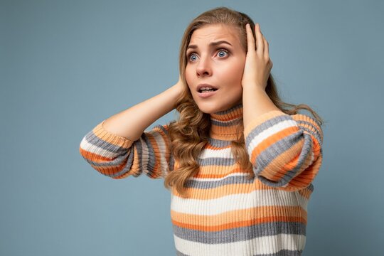 Young shocked pretty nice blonde curly woman with sincere emotions wearing casual striped pullover isolated on blue background with free space and covering ears trying not to hear