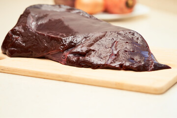 A piece of beef liver on a chopping board. In the background, a dish of vegetables. Selective focus.
