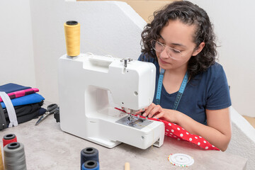 portrait of attractive latin woman using sewing machine, in addition to scissors, threads, fabric, making clothes at home as little businesswoman