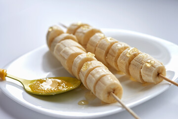 Bananas, strung on sticks, lie on a white plate. Next to them is a spoon of honey. Selective focus.