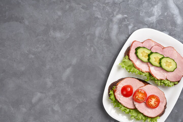 Two sandwiches with ham, cucumber and cherry tomato on a white plate on a gray background. Copy space.