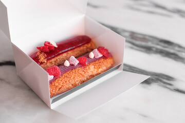 Two eclairs in a white take away box. Dessert with raspberry, jam, marshmallow, cream on a marble background