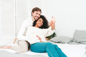 Smiling beautiful woman and her handsome boyfriend. Happy cheerful multiracial family taking selfie. Multiethnic models lying in bed and hugging in white interior. Embracing each other