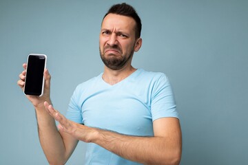 Dissatisfied handsome young unshaven brunet man wearing everyday blue t-shirt isolated over blue background holding and showing mobile phone with empty display for cutout looking at camera