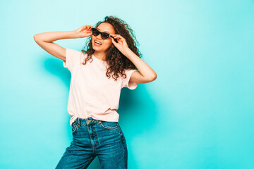 Beautiful black woman with afro curls hairstyle.Smiling model in white trendy t-shirt and jeans clothes. Sexy carefree female posing near blue in studio in sunglasses. Tanned and cheerful