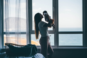 Young mother with long loose flowing hair wanders holding baby in arms near large plastic panoramic windows with sea outside at home.