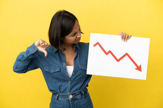 Young mixed race woman isolated on yellow background holding a sign with a decreasing statistics arrow symbol and doing bad signal