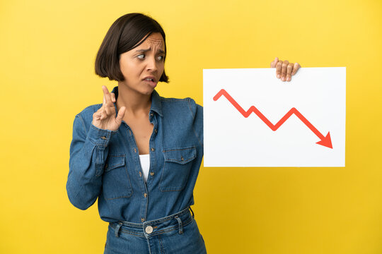 Young mixed race woman isolated on yellow background holding a sign with a decreasing statistics arrow symbol with fingers crossing