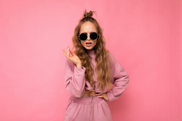Fascinating dissatisfied adult blonde curly woman isolated over pink background wall wearing casual pink sport clothes and stylish sunglasses looking at camera