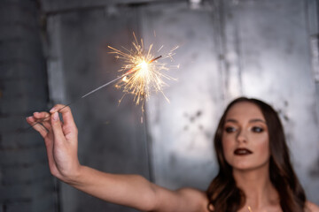 Obraz premium Blurred Beautiful girl, holds bright sparklers in focus in hands. With professional evening make-up, in a gold top, against a gray metal wall with green monsteras background. Holiday party. Copy paste