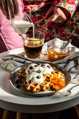 Weekend breakfast. Homemade Belgian waffles with natural whipped cream, chocolate droplets and apricot jam. Woman in flower pajamas pouring milking glass with coffee.  