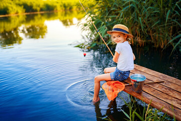 Little boy in straw hat sitting on the edge of a wooden dock and fishing in lake at sunset.