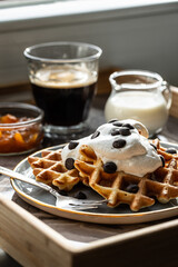 Homemade Belgian waffles with natural whipped cream, chocolate droplets and apricot jam,  glass of coffee , milk jug and jam on marble tray.