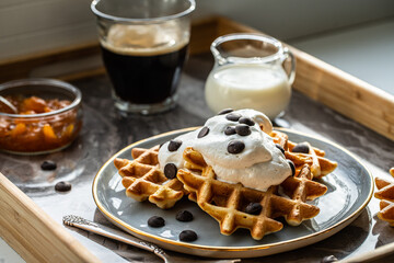 Homemade Belgian waffles with natural whipped cream, chocolate droplets and apricot jam,  glass of coffee , milk jug and jam on marble tray.