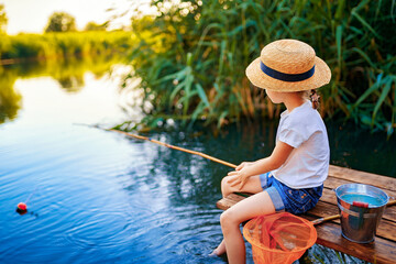 Little boy in straw hat sitting on the edge of a wooden dock and fishing in lake at sunset.