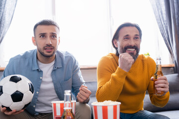 Worried interracial father and son watching football match with beer and popcorn at home