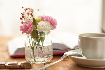 A view of a workplace with pink and white flowers in a small glass jar of water, as well as a red notebook with a pen and coffee on a wooden table. Close-up.