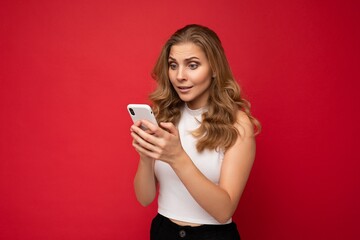 Amazed shocked beautiful young blonde woman wearing white t-shirt isolated over red background using smartphone and texting message via mobile phone looking at gadjet screen