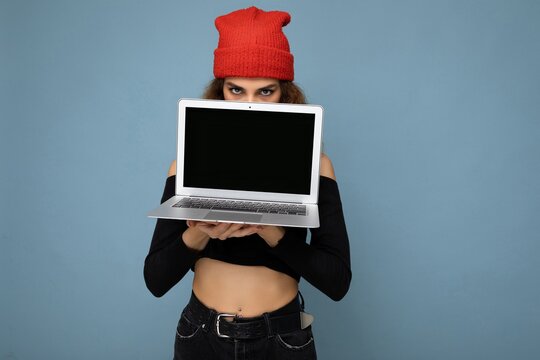 Close-up portrait of funny beautiful dark blond woman holding laptop computer looking at camera wearing black crop top and red and orange do-rag isolated over light blue wall background