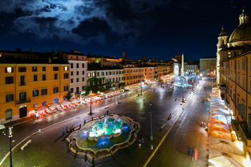 Evening view of an illuminated Piazza Navona with the Neptune and Fountain of Four Rivers lit up as tourists dine at cafes.