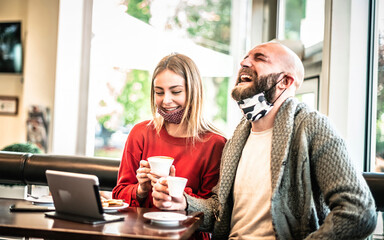 Two happy friends wearing protective face mask having fun together at bar - New normal lifestyle concept with young people on positive mood drinking coffee while working on smart work