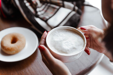 cup of cappuccino with foam in women's hands on the background of the window, coffee