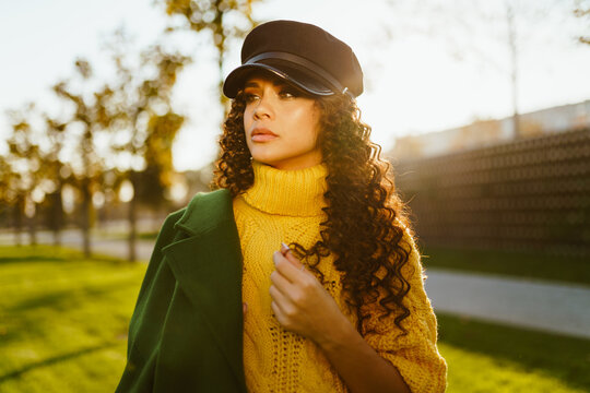 In A Half-turn Turning Her Head With A Cap On Dark Curly Hair Stands In A Bright Yellow Sweater The Girl Is Slapping A Green Coat On Her Shoulder In An Autumn Park. High Quality Photo