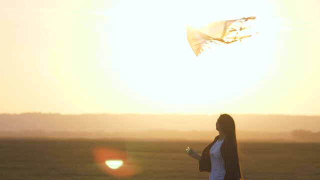 Girl plays with multi-colored kite on field in rays of sun. Teenager dreams of flying and becoming pilot. colored kite hangs in air. Kite Festival. Child and flying kite at sunset in summer.