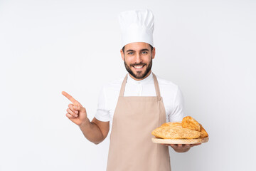 Male baker holding a table with several breads isolated on white background pointing finger to the side