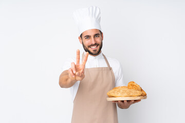 Male baker holding a table with several breads isolated on white background smiling and showing victory sign