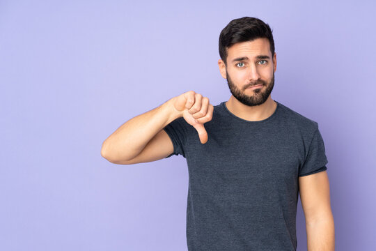 Caucasian handsome man showing thumb down with negative expression over isolated purple background