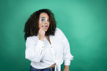 Young african american girl wearing white shirt over green background surprised looking through a magnifying glass