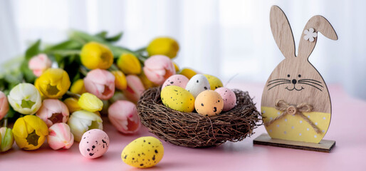 Easter composition with colorful quail eggs in nest, wooden bunny and tulips on pink background. Spring composition. Happy easter greeting card. Banner