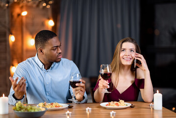 Unhappy Black Man Looking At Girlfriend Talking On Cellphone During Date In Restaurant